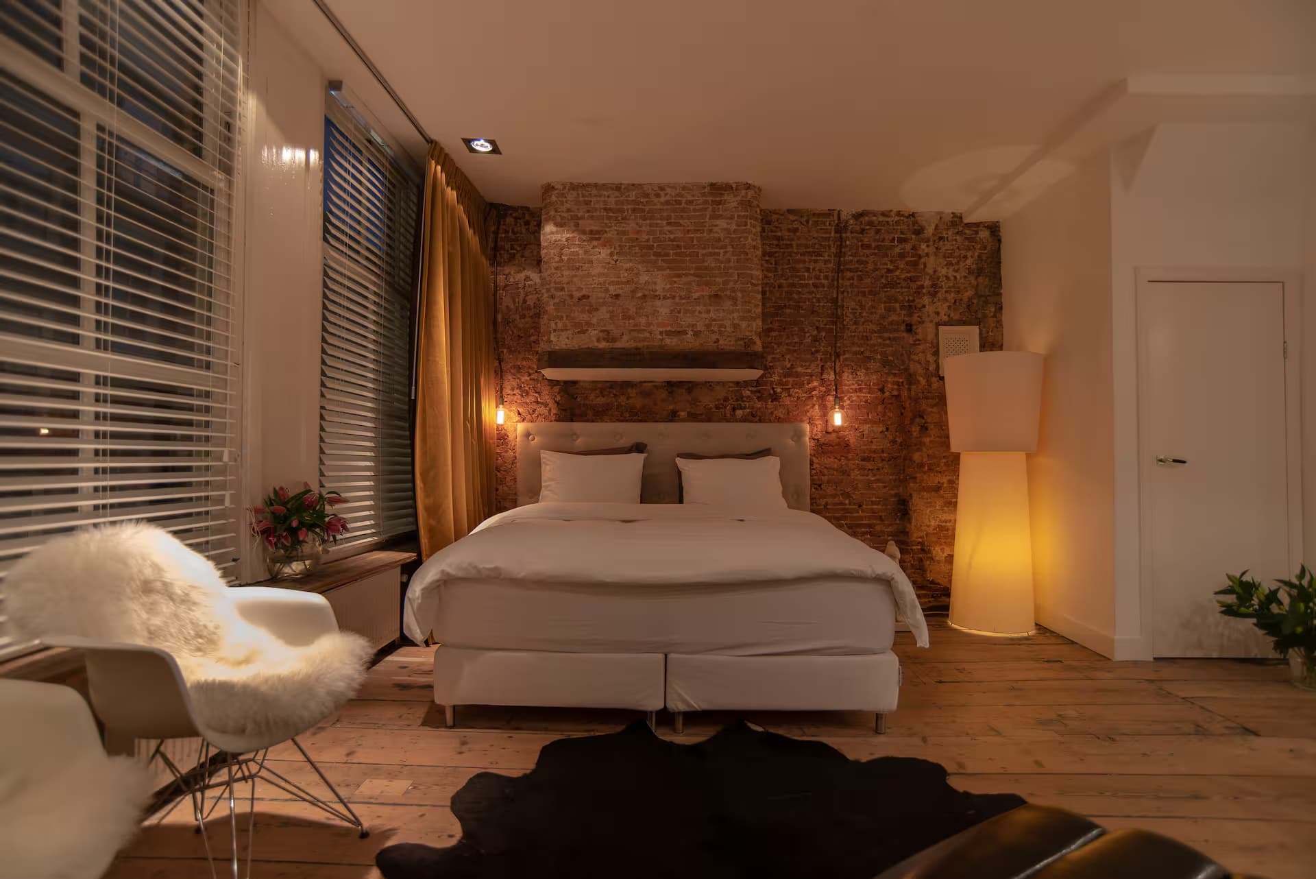 Double Fun — bed in avondlicht met gloeilampen aan kabels en witte fauteuil bij raam met louvre-zonwering