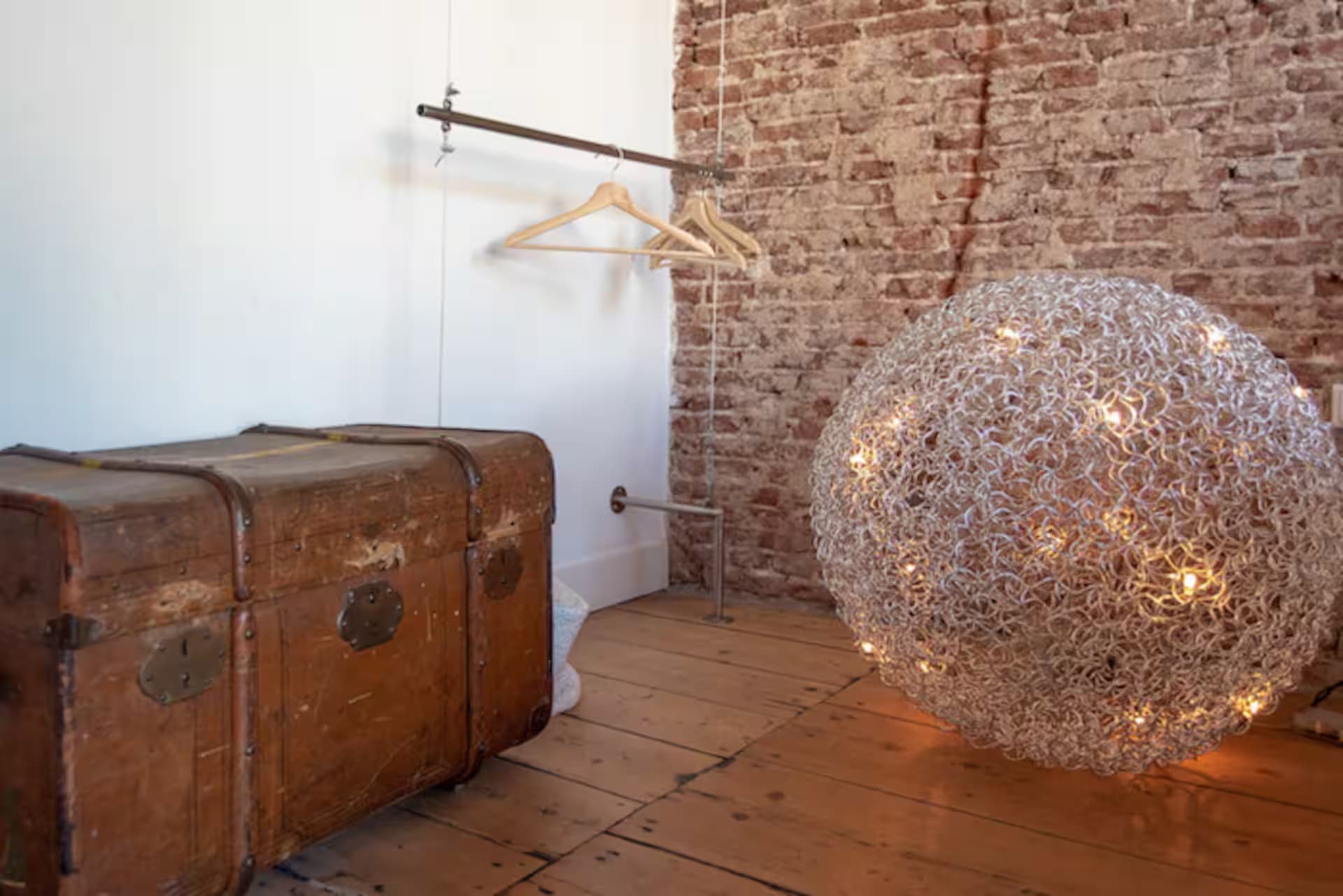 Love Suite — kledingrek-hoek met houten hangers, antieke koffer en gevlochten metalen draadlamp tegen brick-wall